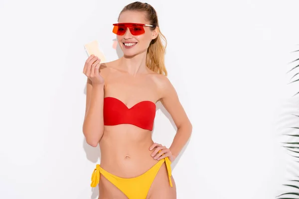 Fröhliche Frau in Badeanzug und Sonnenbrille, die Eis hält, während sie mit der Hand auf der Hüfte auf Weiß steht — Stockfoto