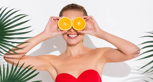 Веселая женщина в купальнике закрывает глаза половинками свежего апельсина на белом — стоковое фото