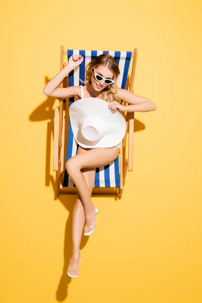 Top view of smiling woman in sunglasses resting in deck chair with straw hat on yellow — Stock Photo