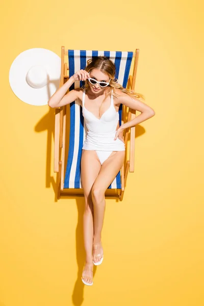 Vista superior de la mujer en traje de baño ajustando las gafas de sol mientras está sentado en la silla de cubierta con la mano en la cadera en amarillo - foto de stock