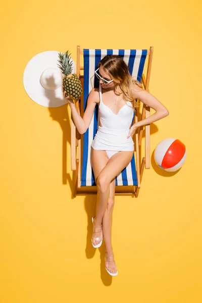 Top view of woman with hand on hip holding fresh pineapple while relaxing in deck chair near straw hat and inflatable ball on yellow — Stock Photo