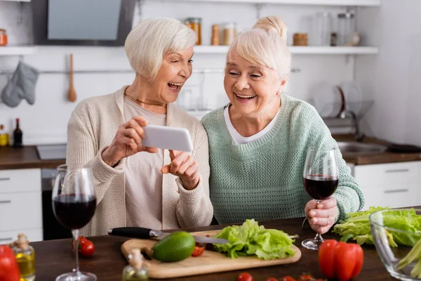 Alegre mujer mayor señalando con el dedo en el teléfono inteligente cerca de amigo y verduras en la mesa - foto de stock