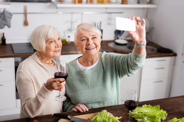 Alegre anciana sosteniendo copa con vino tinto y mirando el teléfono inteligente mientras su amigo se toma selfie en la cocina - foto de stock