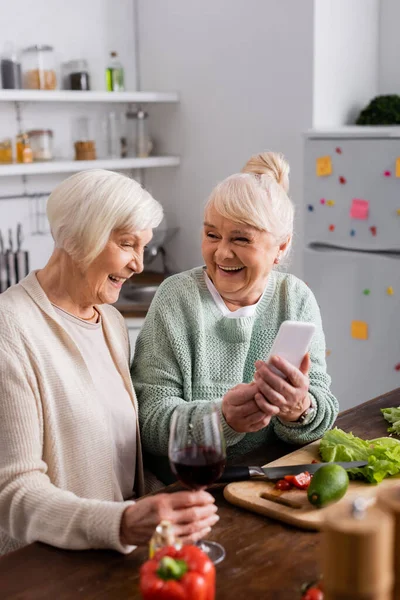 Alegre anciana sosteniendo teléfono inteligente cerca de amigo jubilado en la cocina - foto de stock