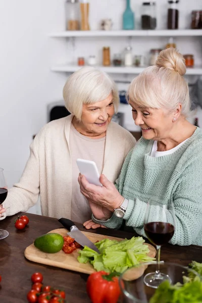 Mulher sênior feliz segurando smartphone perto de amigo aposentado na cozinha — Fotografia de Stock