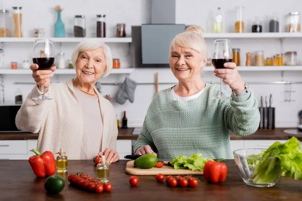 Felices mujeres mayores sosteniendo vasos de vino tinto cerca de verduras frescas en la cocina - foto de stock