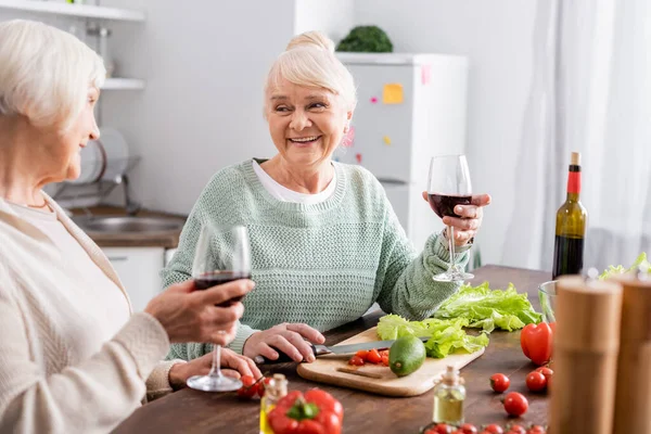 Mulheres sênior sorrindo segurando copos de vinho tinto perto de legumes frescos na mesa — Fotografia de Stock