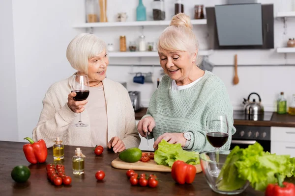 Щаслива старша жінка тримає ніж біля свіжих овочів і подруга на пенсії зі склянкою вина — стокове фото