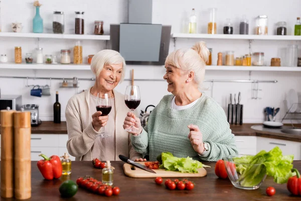 Donne in pensione sorridenti che tengono bicchieri di vino rosso e si guardano vicino a verdure fresche sul tavolo — Foto stock