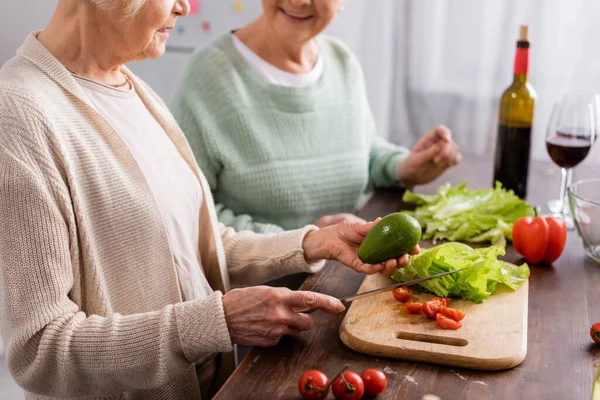Vista ritagliata di donna anziana in possesso di coltello e avocado vicino amico in pensione in cucina — Foto stock
