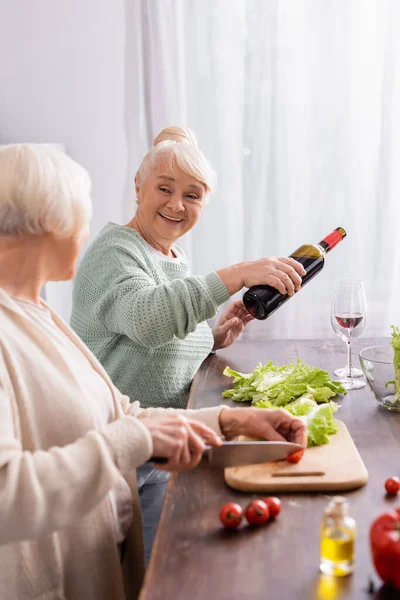 Feliz mujer mayor vertiendo vino tinto en vidrio y mirando a un amigo retirado cortando tomate cherry en primer plano borroso - foto de stock