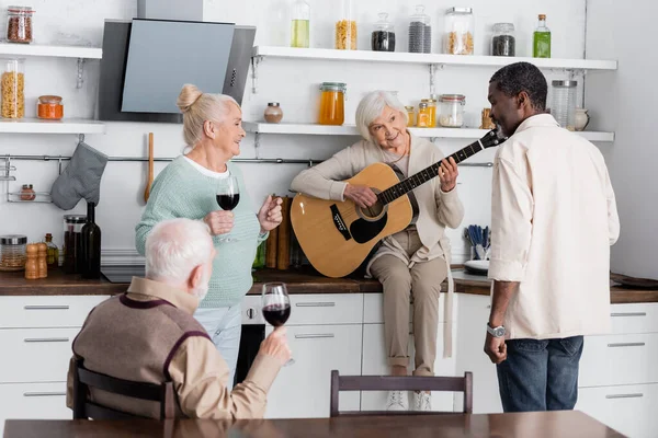 Allegra donna in pensione che suona la chitarra acustica vicino agli amici multiculturali anziani con bicchieri di vino in cucina — Foto stock