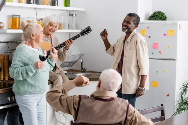 Felice donna in pensione suonare la chitarra acustica vicino allegri amici multiculturali in cucina — Foto stock