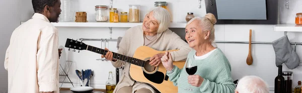 Feliz mujer jubilada tocando la guitarra acústica cerca de amigos mayores multiculturales en la cocina, pancarta - foto de stock