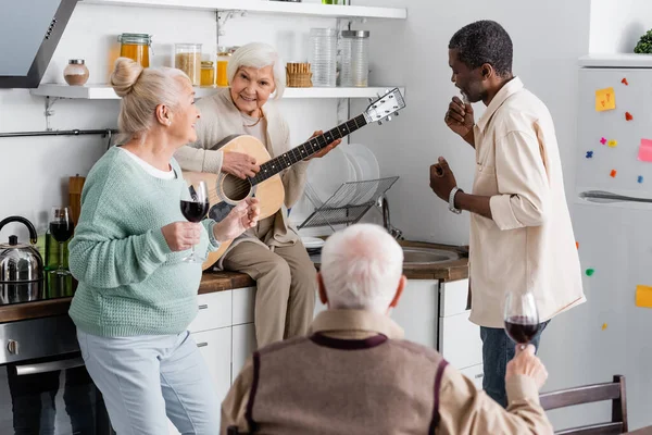 Mulher sênior feliz tocando guitarra acústica perto de amigos multiculturais aposentados na cozinha — Fotografia de Stock