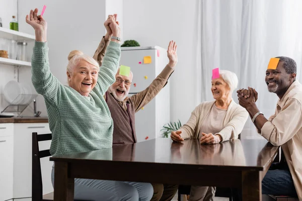 Amigos seniores excitados e multiculturais com notas pegajosas na testa jogando jogo na cozinha — Fotografia de Stock