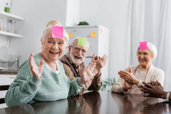 Amigos mayores emocionados y multiculturales con notas adhesivas en la frente jugando adivina quién juego en la cocina - foto de stock