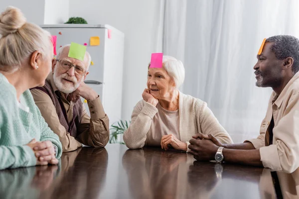 Amigos mayores multiculturales con notas adhesivas en la frente jugando juego en la cocina - foto de stock