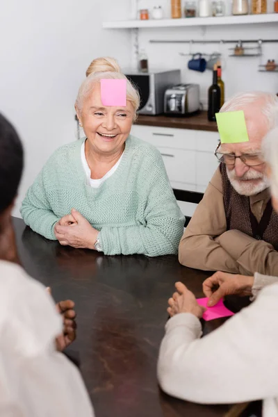 Amigos mayores felices y multiculturales con notas adhesivas en la frente jugando juego en la cocina - foto de stock