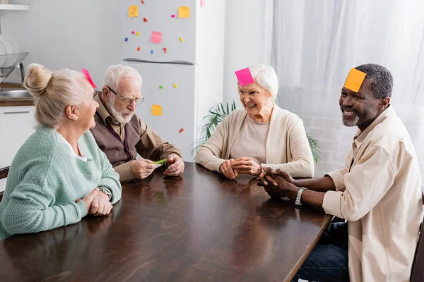 Sorrindo amigos seniores multiculturais com notas pegajosas coloridas na testa jogando jogo na cozinha — Fotografia de Stock