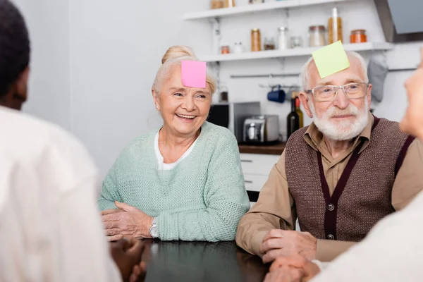 Sonriente hombre y mujer senior con notas pegajosas coloridas en la frente jugando juego con amigos multiculturales en primer plano borroso - foto de stock