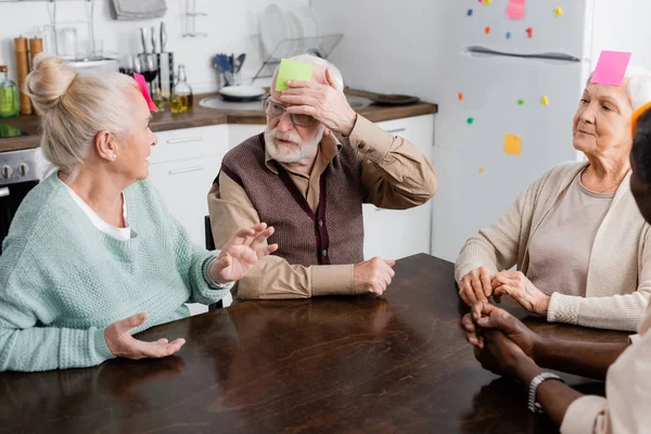 Amigos mayores multiculturales con notas pegajosas coloridas en la frente jugando juego en la cocina - foto de stock