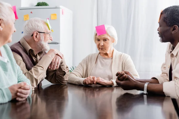 Pensive senior woman with sticky note on forehead playing game with multicultural friends in kitchen — Stock Photo