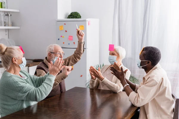 Excited multicultural senior friends in medical masks with sticky notes on foreheads applauding while playing game in kitchen — Stock Photo