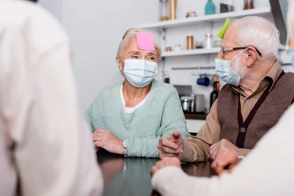 Senior hombre y mujer en máscaras médicas con notas adhesivas en la frente jugando juego con amigos multiculturales en primer plano borrosa - foto de stock