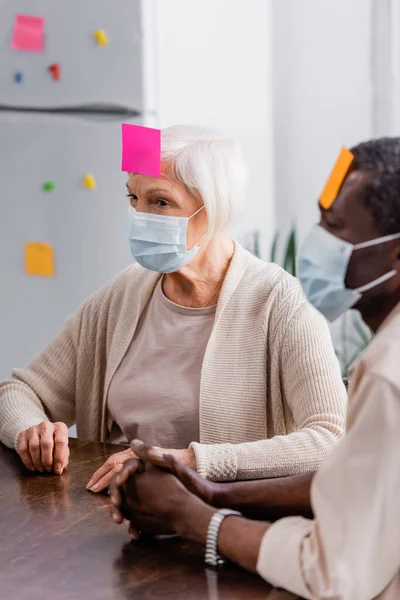 Multicultural senior friends in medical masks with sticky notes on foreheads playing game in kitchen — Stock Photo