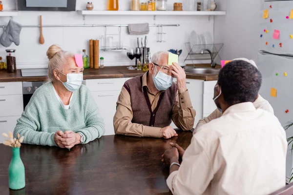 Retired man and woman in medical masks with sticky notes on foreheads playing game with multicultural friends — Stock Photo