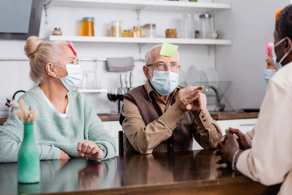 Personas jubiladas en máscaras médicas con notas adhesivas en la frente jugando a juegos con amigos multiculturales - foto de stock