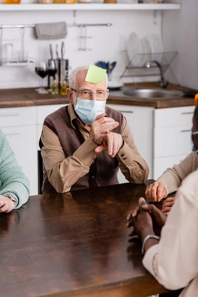 Hombre retirado en máscara médica con nota adhesiva en la frente jugando juego con amigos multiculturales - foto de stock