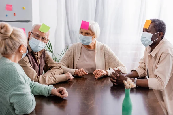 Retired people in medical masks with sticky notes on foreheads playing game with interracial friends — Stock Photo