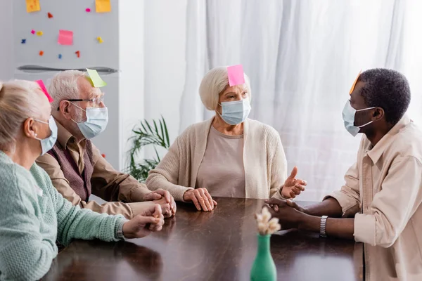 Retired people in medical masks with sticky notes on foreheads discussing while playing game with multiethnic friends — Stock Photo