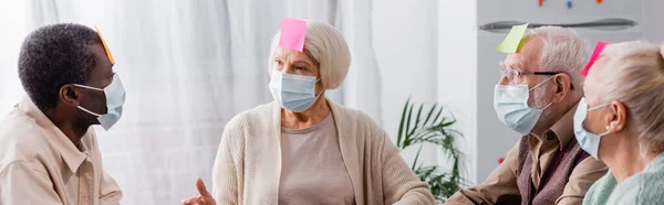 Retired people in medical masks with sticky notes on foreheads discussing while playing game with multiethnic friends, banner — Stock Photo