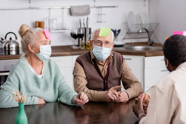 Retired man in medical mask with sticky note on forehead looking at camera while playing game with multiethnic friends — Stock Photo