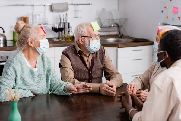 Retired multiethnic people in medical masks with sticky notes on foreheads playing game at home — Stock Photo