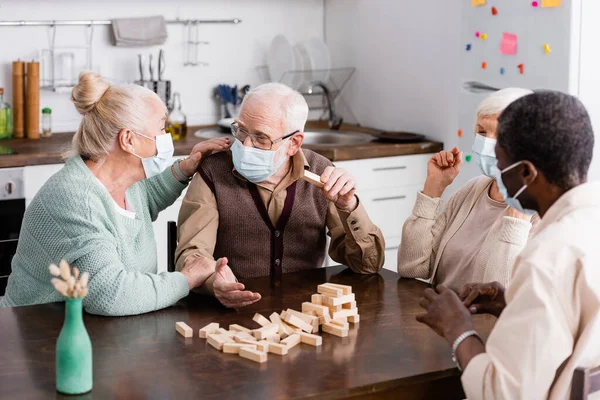 Personas jubiladas multiculturales en máscaras médicas jugando bloques de madera torre juego en casa - foto de stock