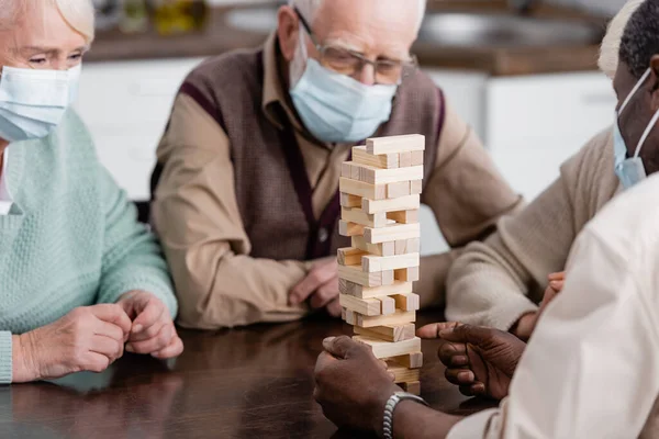 Pensionato uomo africano americano in maschera medica giocare torre di legno blocchi di gioco vicino amici su sfondo sfocato — Foto stock