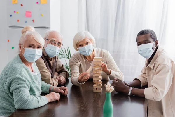Pensionati interrazziale persone in maschere mediche giocare torre di legno blocchi di gioco a casa — Foto stock