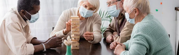 Personas jubiladas multiculturales en máscaras médicas jugando bloques de madera torre juego en casa, pancarta - foto de stock