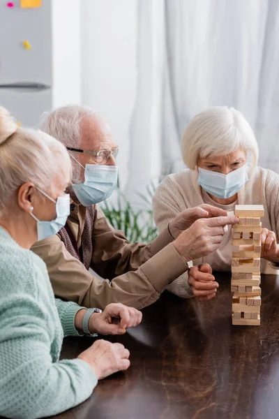 Amigos aposentados em máscaras médicas jogando torre blocos de madeira jogo em casa — Fotografia de Stock