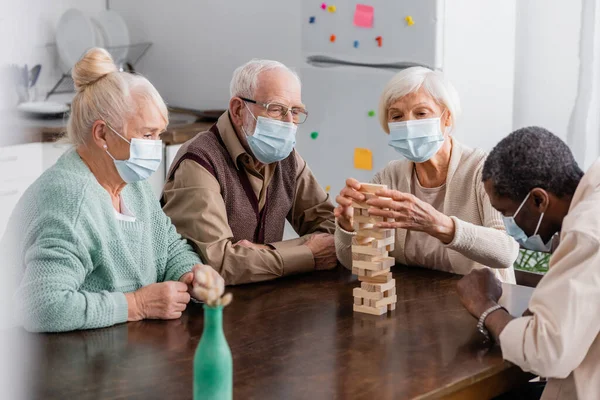 Aposentados amigos inter-raciais em máscaras médicas jogando torre blocos de madeira jogo em casa — Fotografia de Stock