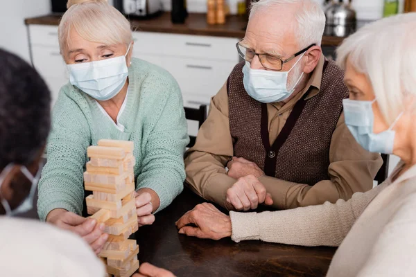 Retired people in medical masks playing tower wood blocks game near african american friend on blurred foreground — Stock Photo