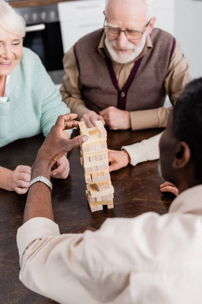 Homme afro-américain à la retraite jouant tour blocs de bois jeu près heureux amis aînés sur fond flou — Photo de stock