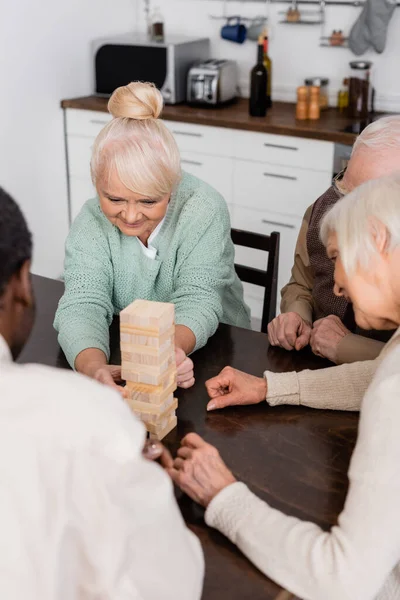 Aposentados multiculturais alegres jogando jogo de blocos de madeira de torre em casa — Fotografia de Stock