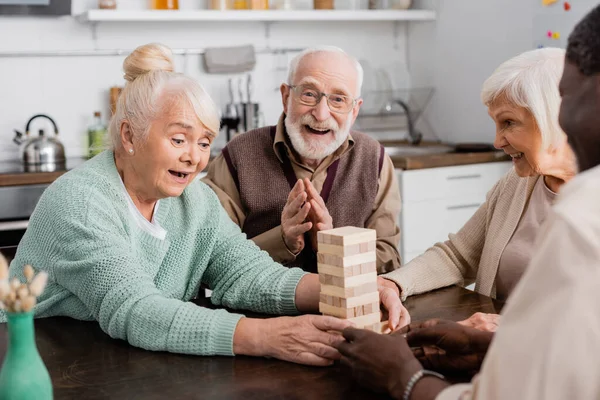 Stupito donna in pensione giocando torre di legno blocchi di gioco vicino allegri amici multiculturali — Foto stock