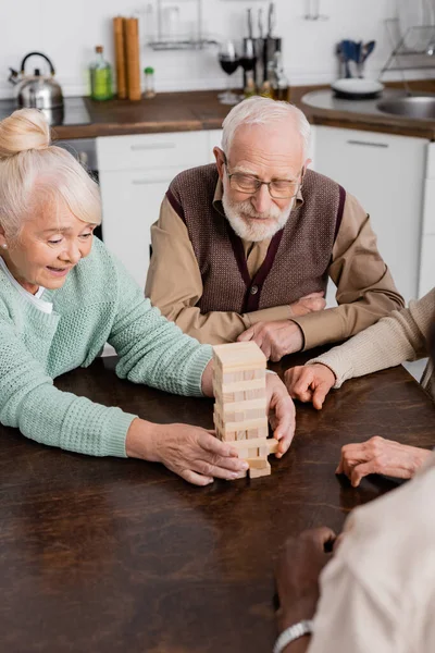 Amigos mayores multiculturales felices jugando bloques de madera torre juego en casa - foto de stock