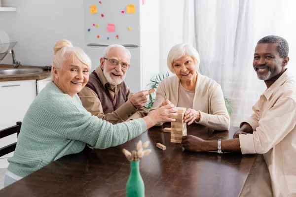 Happy interracial pensioners looking at camera while playing tower wood blocks game at home — Stock Photo